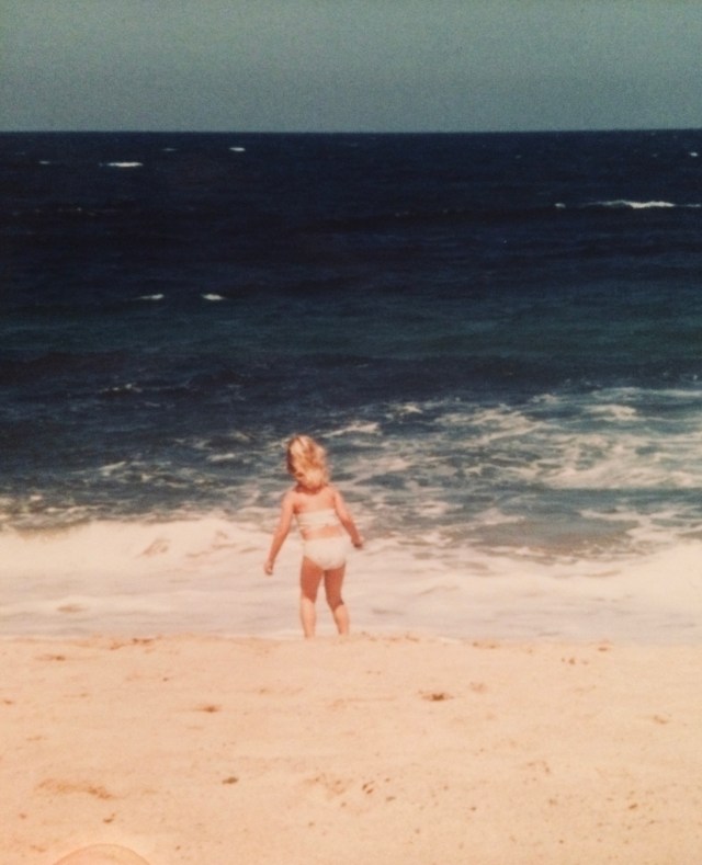 A young girl stands in front of crashing waves on a beach with her back to the camera.