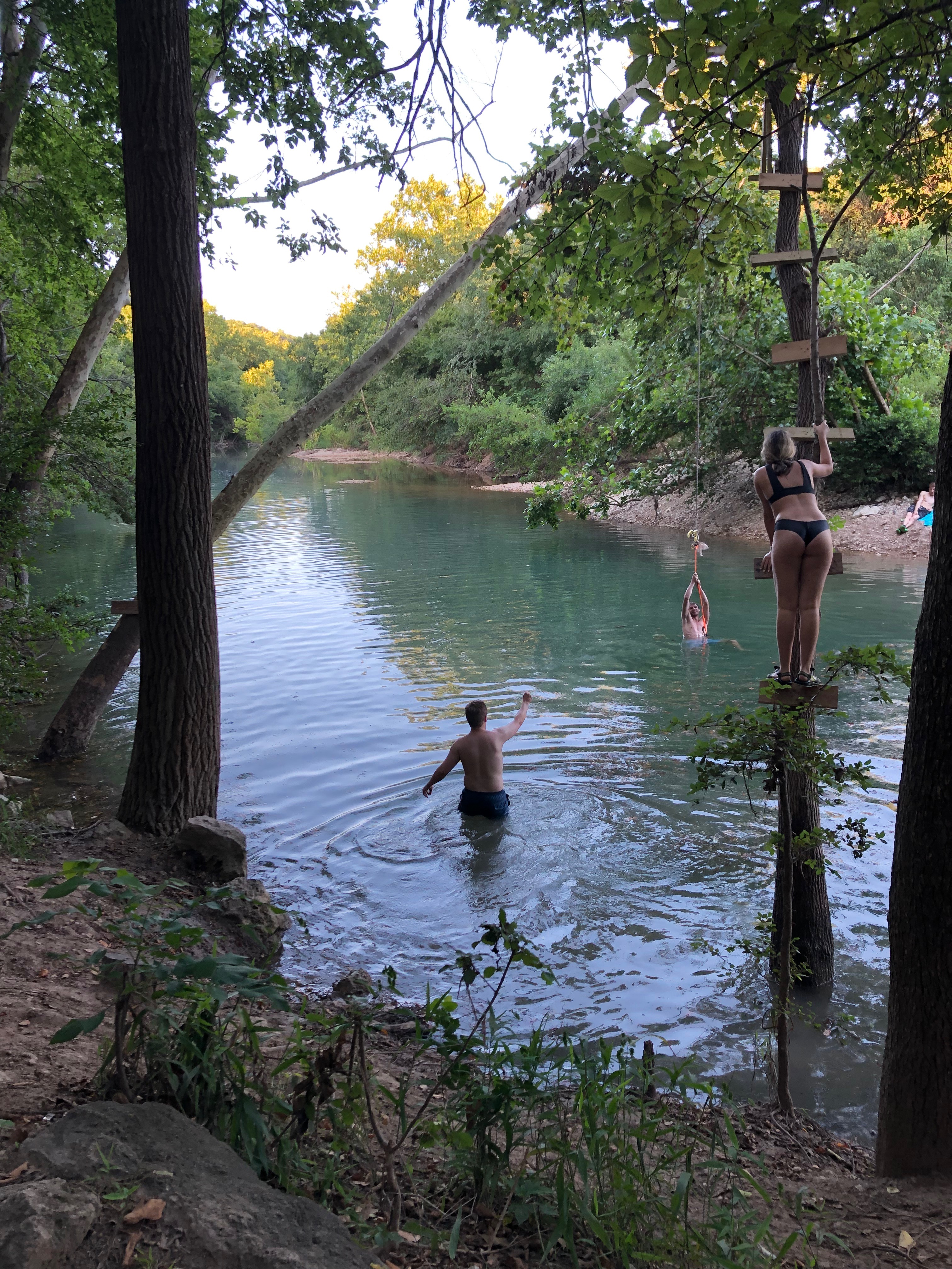 Girl climbing tree over water waits to receive rope from friend to swing.