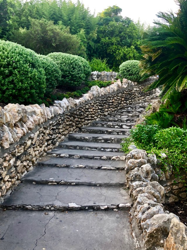 Stairway lined by stacked rocks and surrounded by trimmed bushes.