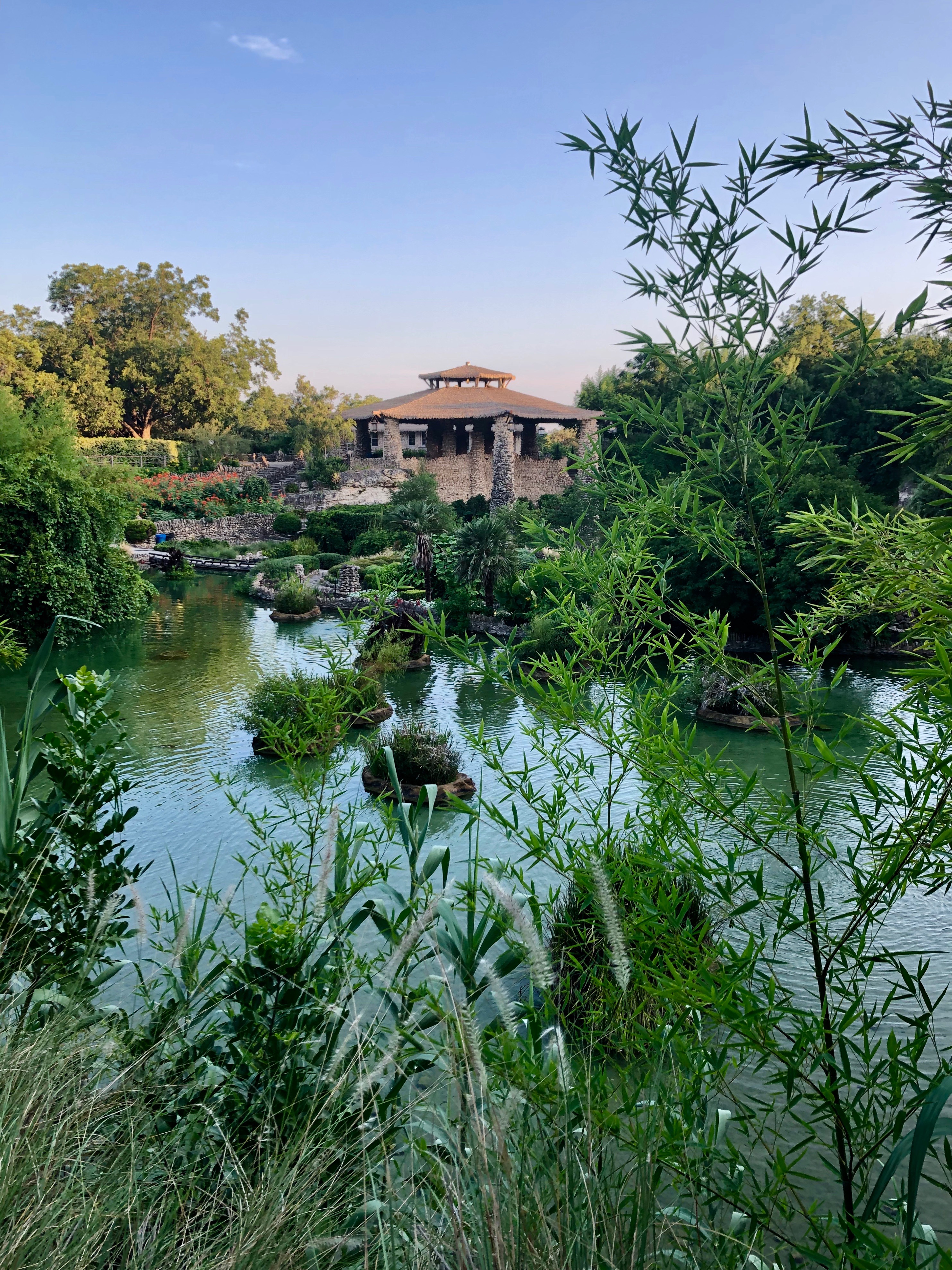Large, stacked stone pavilion can be seen from across the pond and in between bushes and bamboo.