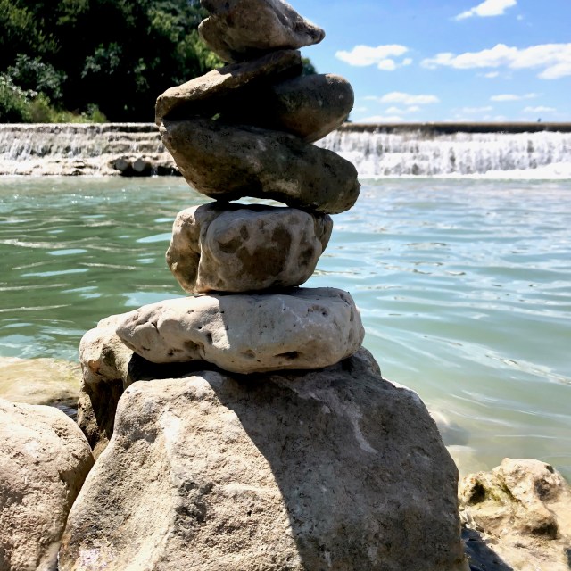 Six large rocks are stacked on a bigger boulder that sits in the green river below the dam.