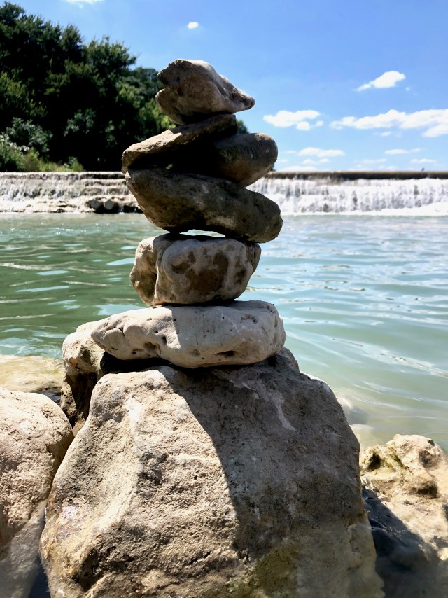 Six large rocks are stacked on a bigger boulder that sits in the green river below the dam.